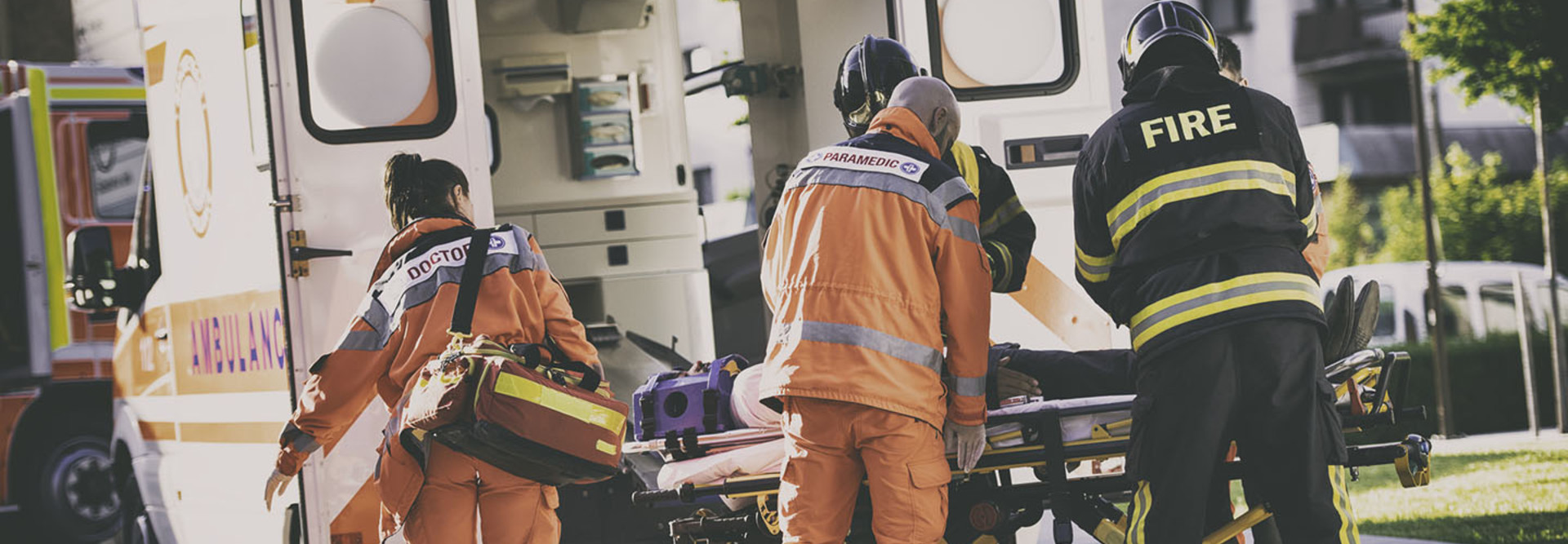 Two firefighters helping one female and one male paramedic to put a stretcher with injured person into ambulance vehicle.