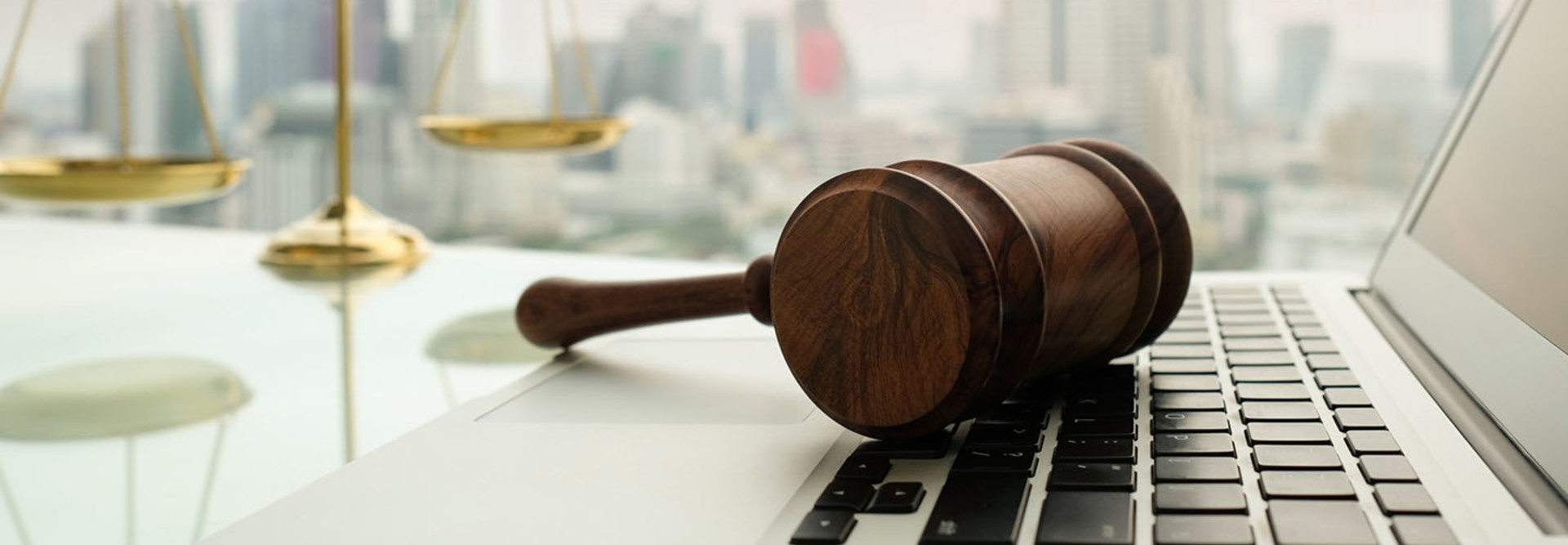 Wooden gavel on laptop keyboard with scales of justice in business city background.