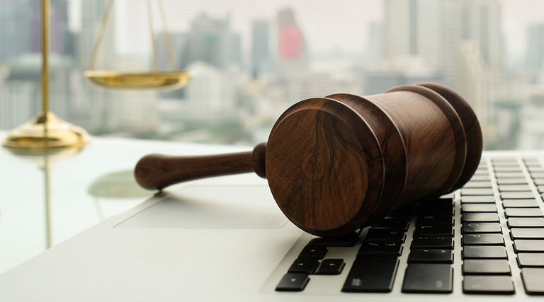 Wooden gavel on laptop keyboard with scales of justice in business city background.