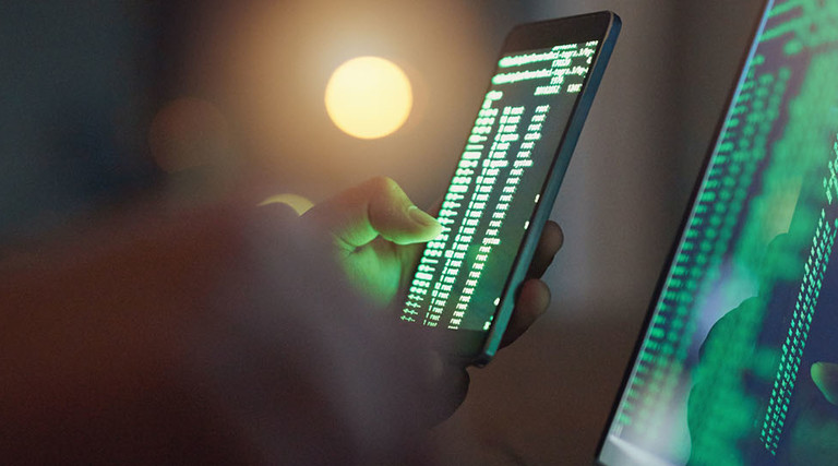 Shot of an unrecognizable hacker using a cellphone and laptop in the dark